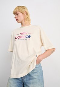 Personne blonde portant un t-shirt ample crème de New Balance avec un texte rose et bleu, un jean bleu clair, se tenant devant un fond blanc uni.
