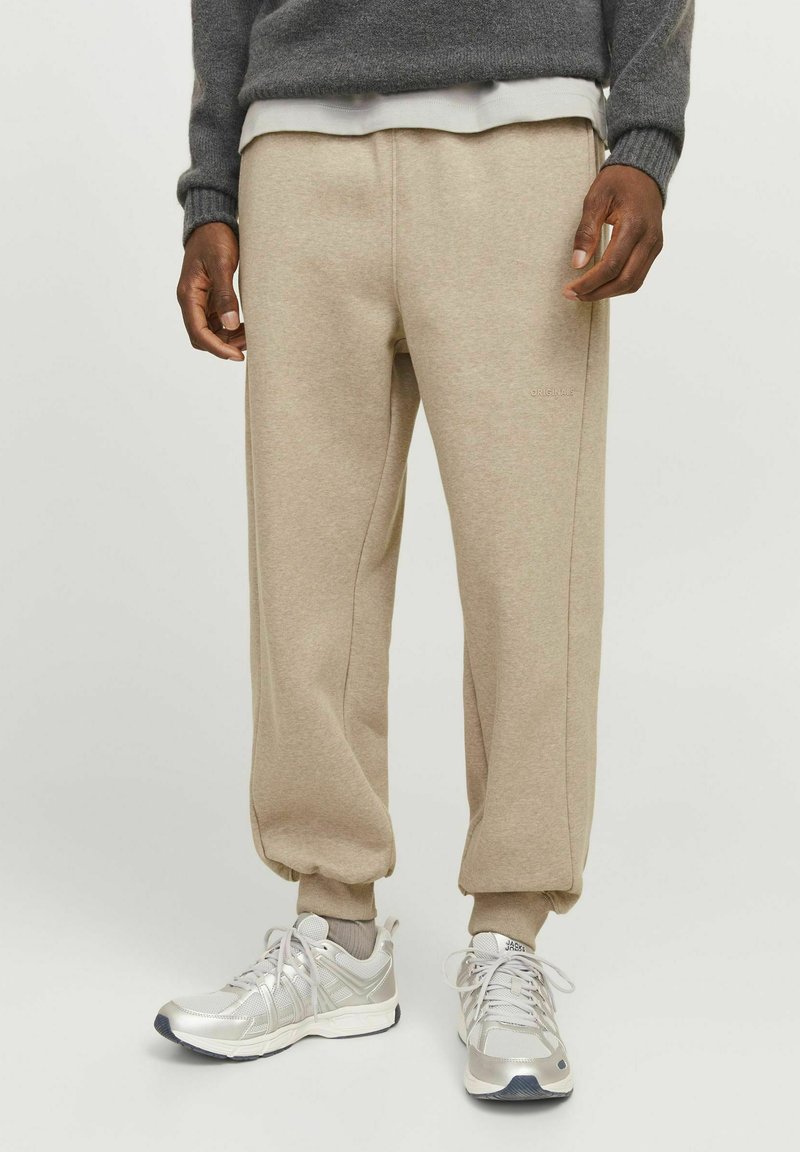 Beige tapered sweatpants med elastiske manchetter, der har en afdæmpet tekstur og detaljer langs sidesømmene, parret med sølvfarvede sneakers.