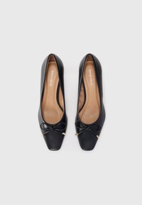 Black leather flats with a square toe, featuring a small bow detail and gold hardware accents. Soft brown interior lining visible.