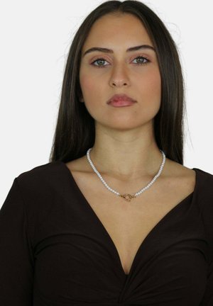 Collana di perle con piccole perle bianche e un fermo circolare in oro, indossata da un modello con un top nero a V su uno sfondo bianco.