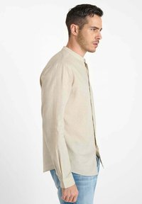Man die naar rechts kijkt, gekleed in een beige lange mouw mandarin kraag shirt en lichtblauwe jeans tegen een witte achtergrond.