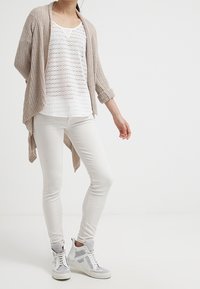 Beige gestrickte Strickjacke, weißes Spitzenmuster-Top, weiße Slim-Fit-Hose und graue High-Top-Sneaker mit weißen Akzenten. Lässiges, geschichtetes Outfit.