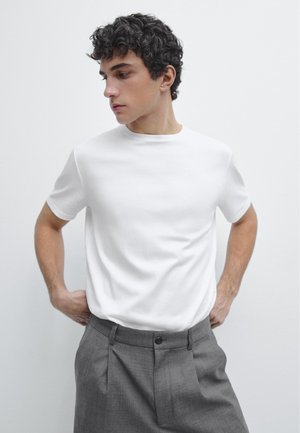 Jonge man met krullend haar, gekleed in een eenvoudig wit T-shirt dat in hoogwatergrijze broek is gestopt, kijkt naar links met zijn handen achter zijn rug.