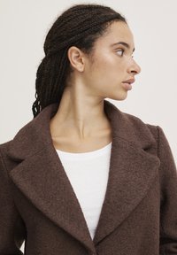 Manteau en laine texturée marron avec un col châle, associé à un t-shirt blanc à col rond. Le mannequin affiche le profil latéral du manteau.