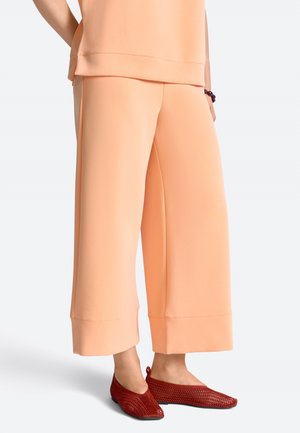 Dolna część ciała ubrana w brzoskwiniowe, szerokie, skrócone spodnie z szerokimi nogawkami i czerwone plecione płaskie buty, z jedną ręką spoczywającą na biodrze.