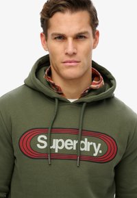 Oliwkowa bluza z kapturem z szarymi sznurkami i czerwono-czarnym logo "Superdry" z przodu. Pod spodem znajduje się warstwowa koszula w kratę.