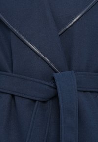 Manteau en laine bleu marine avec une ceinture en tissu assortie nouée à la taille et des revers en cuir qui se croisent à l'avant.