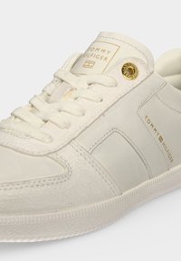 Λευκό sneaker Tommy Hilfiger με υφές σουέτ και δέρματος, χρυσή ετικέτα με λογότυπο στη γλώσσα και χρυσή λεπτομέρεια οφθαλμών κοντά στα κορδόνια.