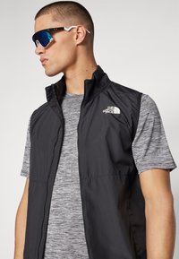 Mustmata ilma varrukatega vest kõrge krae ja tõmblukuga, sobitatud halli lühikeste varrukatega särgiga. Üksikud sinaka varjundiga prillid lõpetavad riietuse.