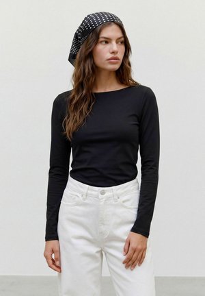 Femme aux longs cheveux bruns portant un haut noir à manches longues, un pantalon blanc et un foulard noir à pois, regardant sur le côté.