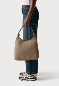 Brun läder axelväska med en rundad design, utrustad med en platt ficka och ett robust handtag, i kombination med blå vida jeans och sneakers.