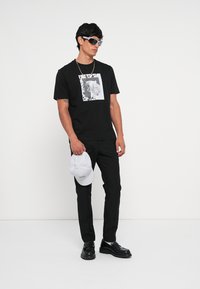 Camiseta negra con un diseño gráfico en el frente, combinada con pantalones negros, gorra blanca y zapatos negros, con gafas de sol como accesorios.