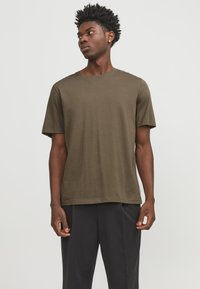 Olive green cotton t-shirt with short sleeves, crew neckline, relaxed fit, and a small label accent at the hem, worn with dark trousers.