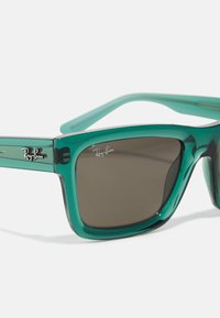 Ochelari de soare din plastic verde translucid, cu lentile de culoare maro închis. Caracteristicile includ un design pătrat și logo-ul Ray-Ban pe brate.