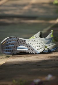 Chaussures de course gris clair et bleu avec des accents jaune néon posées sur une surface extérieure pavée, une semelle visible avec motif de la bande de roulement.