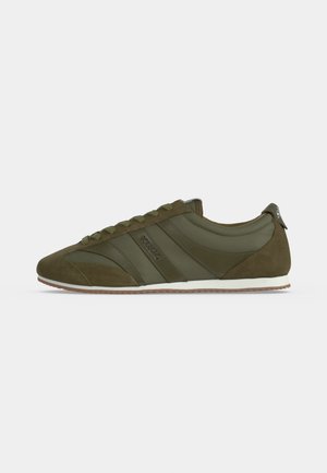Zapatilla deportiva de perfil bajo en color verde oliva con detalles de ante, cierre con cordones, entresuela blanca y suela de goma vista desde un lateral.
