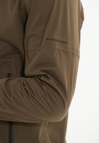 Manche de veste de sport marron avec une texture lisse et légèrement brillante ; arbore le mot "ENDURANCE" imprimé en noir sur le bras.