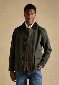 Mann trägt eine olivgrüne Jacke mit schwarzem Kragen, pinkfarbenen Pullover, gelbes Hemd mit Kragen und blaue Jeans vor einem einfarbigen beigen Hintergrund.