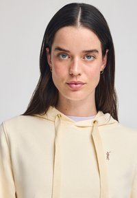 Lysegul hætte sweatshirt med en central kengurulomme, snoretræk og et lille grafisk logo på brystet. Blød tekstur.