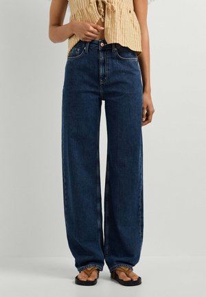Vrouw draagt hooggetailleerde donkerblauwe wijde jeans, tan mouwloos overhemd met knopen en bruine sandalen, staand tegen een lichte achtergrond.