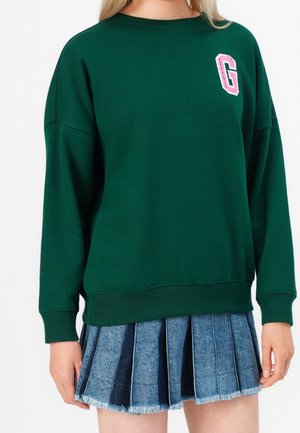 Osoba má na sobě tmavě zelenou mikinu s růžovým logem "G" na hrudi a modrou džínovou plisovanou sukni s třepeným lemem.