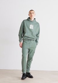 Oliven grønn hettegenser og matchende joggebukser med frontlomme og hvit "HUGO"-logo. Brukt med svarte snørebåndsko. Minimalistisk design.
