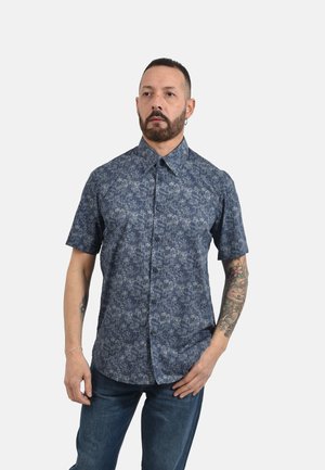 Uomo con capelli corti e tatuaggi sul braccio destro, che indossa una camicia blu a maniche corte con bottone e jeans blu, in piedi davanti a uno sfondo bianco.