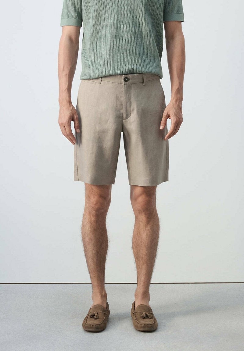 Homme portant un short beige mi-genou, un pull léger vert clair à manches courtes et des mocassins à pampilles marron, debout devant un fond uni.