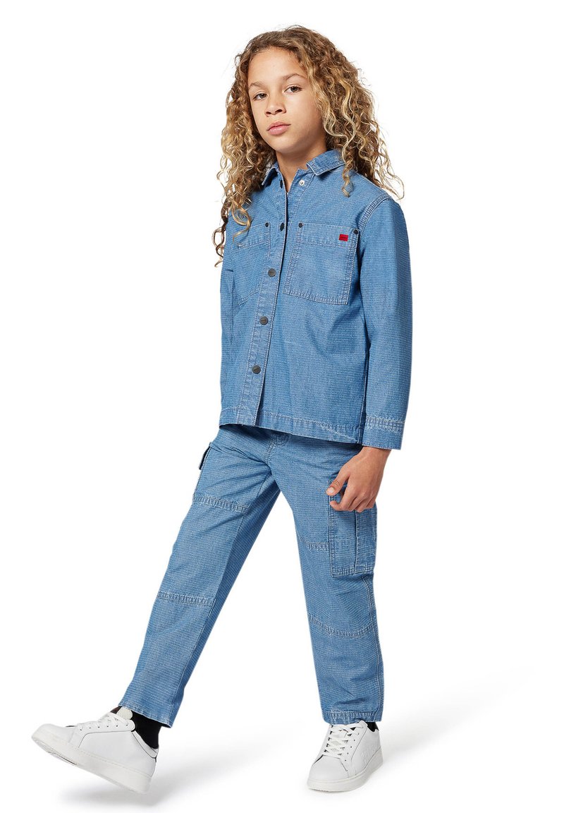 Tenue en denim composée d'une veste boutonnée bleu clair et d'un pantalon assorti, avec des poches avant et des baskets blanches.
