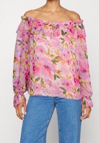 Haut épaules dénudées en tissu rose transparent avec imprimé floral, col à volants et manches longues avec poignets froncés. Associé à un jean bleu.