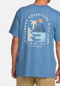 Himmelblå T-skjorte med grafisk design av palmer, fjell og en sol. Logo-teksten leser "Billabong Adventure Division."