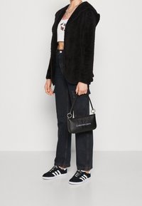 Zwarte, fuzzy hoodie, witte cropped shirt met bloemenborduurwerk, donkere high-waisted jeans, zwarte Calvin Klein handtas en zwarte Adidas sneakers.