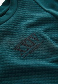 Tyrkysová textúrovaná tkanina s diamantovým vzorom, s tmavo hnedým vyšitým logom, ktoré znie "XXIV SEVEN."