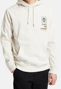 Sudadera color crema hecha de una tela texturizada, con un bolsillo frontal, cordones y un logo impreso con texto en el pecho.