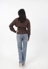 Suéter de punto marrón con cintura acanalada, combinado con jeans claros de campana. La modelo está de pie con la mano en la cadera, vista desde detrás.