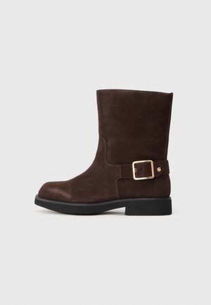Botină din suede maro până la gleznă, cu vârf rotund, talpă neagră plată și accent cu cataramă aurie pe lateral. Textură netedă și design simplu.