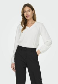Hvid, langærmet bluse med V-hals og elastiske manchetter, stylet med sorte, højttaljede bukser. Glat tekstur, afslappet pasform.