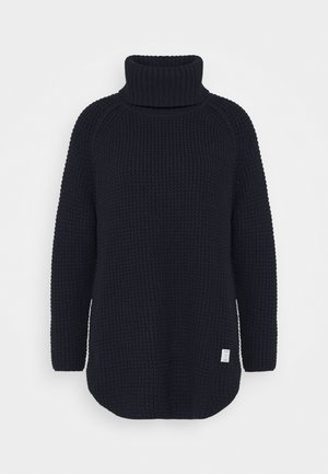 Pull à col roulé en grosse maille noire avec manches longues et petite étiquette blanche sur le bas devant.