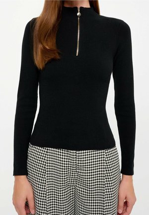 Femme portant un haut noir moulant à manches longues avec une fermeture éclair à l'avant et un pantalon taille haute à motif pied-de-poule noir et blanc.
