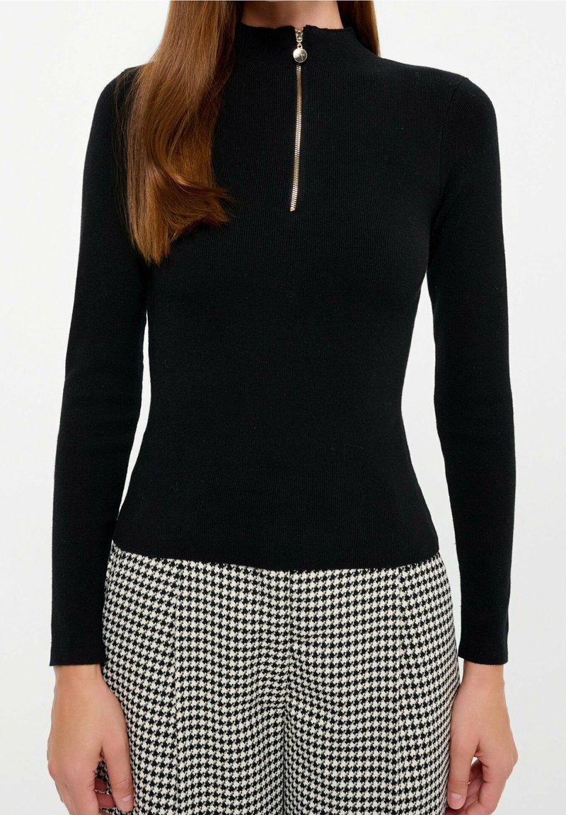 Femme portant un haut noir moulant à manches longues avec une fermeture éclair à l'avant et un pantalon taille haute à motif pied-de-poule noir et blanc.