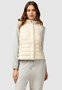 Gevoerde mouwloze vest in romige kleur met horizontale stiksels, hoge kraag en zijzakken. Gedragen over een lichtgrijs langslavend shirt.