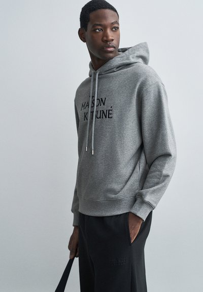 Grijze hoodie met trekkoorden, voorzien van zwarte tekst "MAISON KITUNÉ" op de voorkant. Gecombineerd met zwarte broek met een subtiel logo.