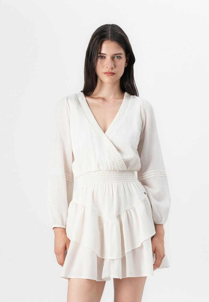 Il modello si posa con sicurezza, indossando un vestito avvolgente color crema con maniche lunghe e una gonna a strati, su uno sfondo bianco semplice.