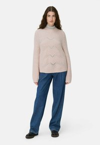 Helles rosa gestricktes Sweater mit einem strukturierten Muster, hohem Kragen und weiten Ärmeln, kombiniert mit dunkelblauen weiten Jeans und schwarzen Schuhen.