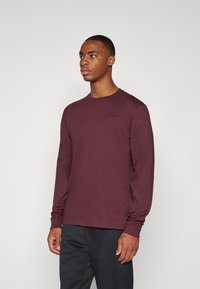Langermet maroon Nike-skjorte med rund hals og en liten logo på brystet, laget av mykt stoff. Kombinert med svarte bukser.