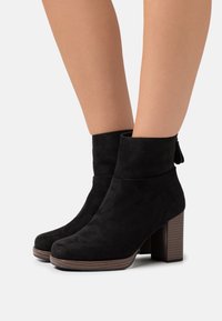 Black suede ankle boots with a block heel and a rounded toe. Features a cuffed top and textured wooden sole with a subtle pattern.