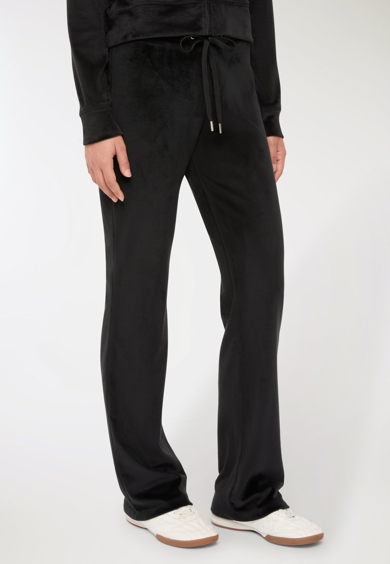 Osoba ubrana w czarne dresowe spodnie z weluru z sznurkiem w talii oraz białe trampki, stoi na tle jednolitego tła.