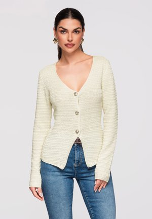 Femme aux cheveux foncés portant un cardigan boutonné en maille crème texturée et un jean bleu, debout devant un fond gris clair uni.