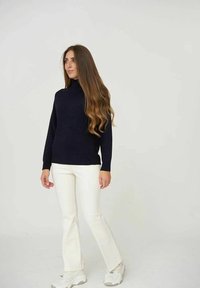 Pullover aderente a collo alto di colore navy abbinato a pantaloni bianchi aderenti e sneakers chiare, in piedi su uno sfondo neutro.