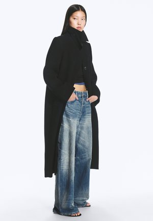 Frau trägt einen langen schwarzen Mantel über einem schwarzen kurzen Oberteil und weit geschnittenen, ausgewaschenen blauen Jeans, steht mit den Händen in den Taschen vor weißem Hintergrund.